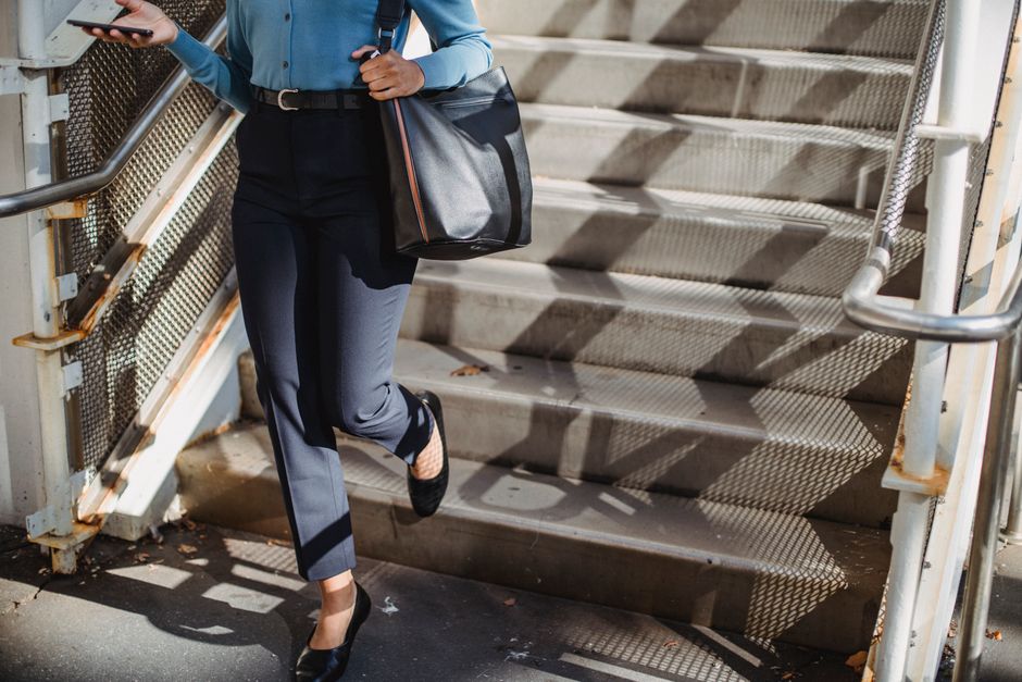 Anonymous female in elegant wear with bag walking down staircase with railing while texting message on cellphone on sunny street