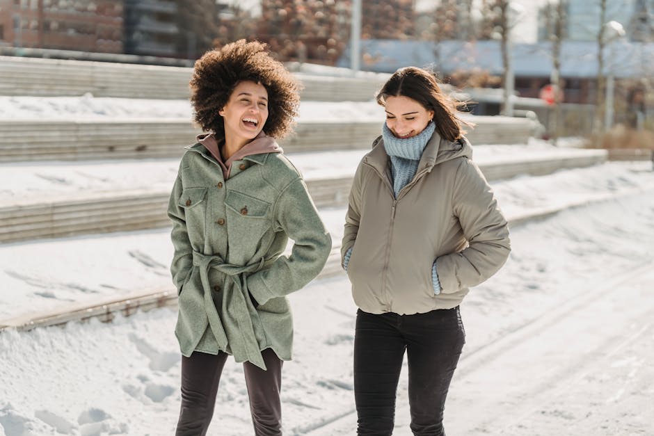 Expressive young multiracial female friends wearing warm clothes chatting and walking together with hands in pockets on snowy city street on freezing winter day