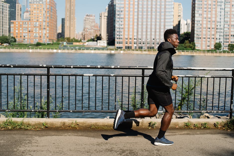 Focused man jogging beside a river with a city skyline in the bright daylight.