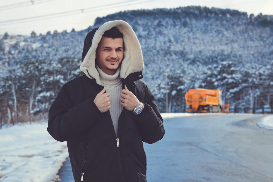 Man wearing a hooded jacket standing on a snowy road with mountains in the background.