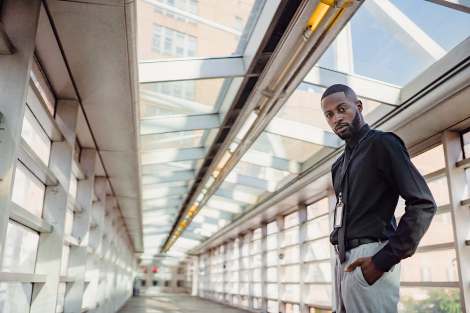 A fashionable man poses confidently in a modern glass corridor with urban architecture.