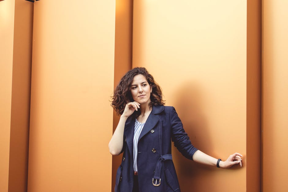 Confident woman in blue coat leaning against a vibrant orange wall, exuding style and elegance.