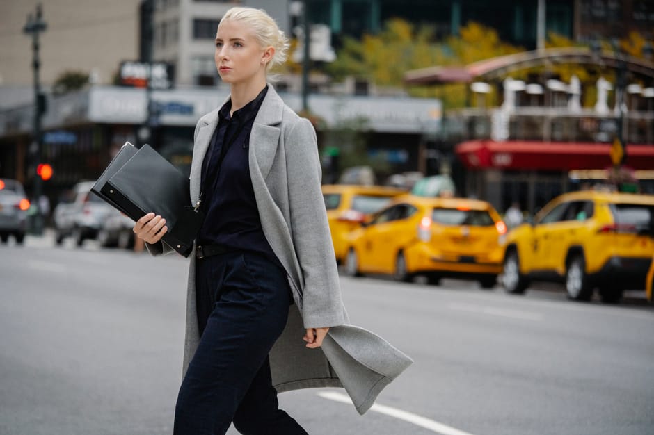 Young confident well dressed blond female entrepreneur with folders walking on asphalt roadway while looking forward in town