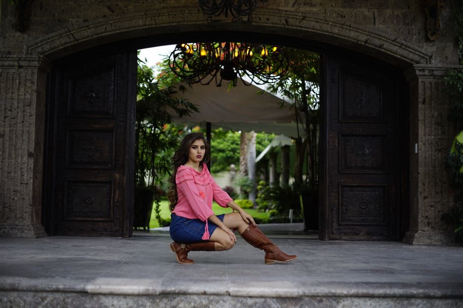 Stylish woman in a pink top and cowboy boots poses confidently outdoors in an elegant setting.