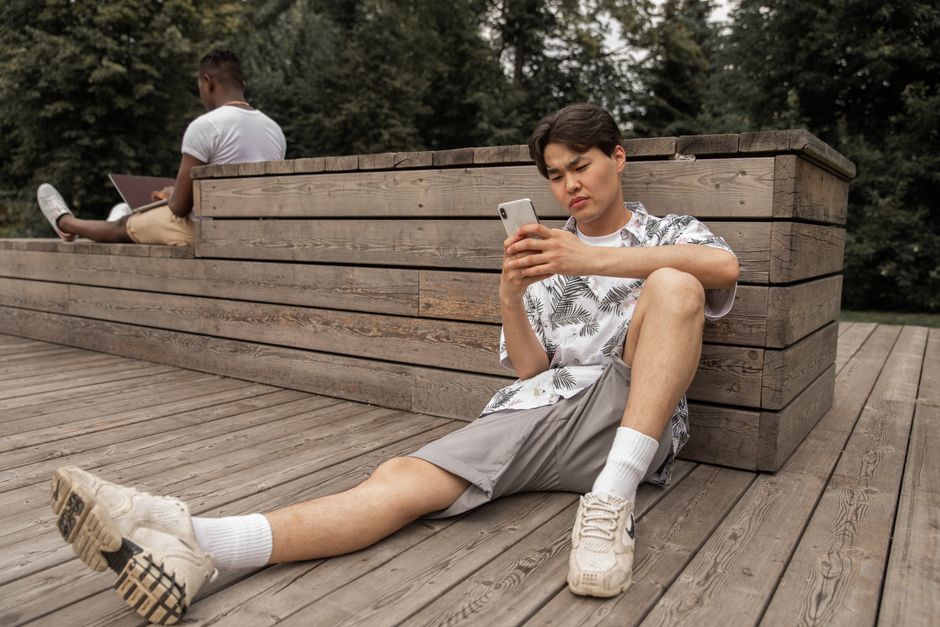 Full body of focused ethnic male in trendy outfit resting on wooden platform in park while messaging on smartphone