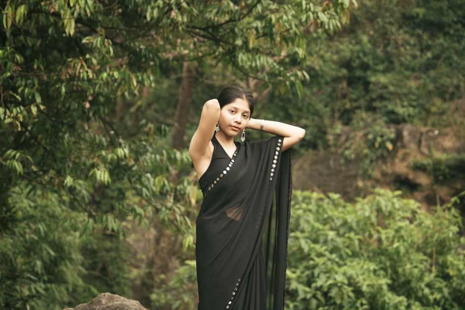 Woman in black saree posing gracefully in lush green outdoor setting.