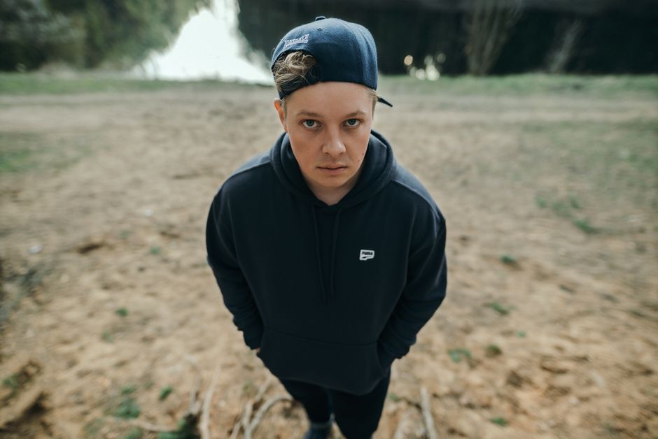 A young man in a casual hoodie and cap, gazing thoughtfully outdoors by water.