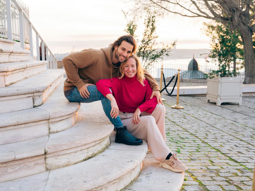 A smiling couple sitting on stone steps in Lisbon, enjoying a warm sunset by the sea.