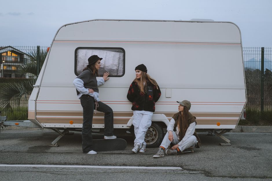 Three young women skateboarders in casual wear hanging out by a camper trailer, enjoying the street lifestyle.