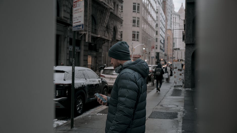 A man checks his phone on a winter day in downtown New York City, surrounded by snowy streets.