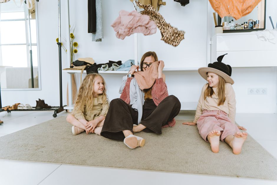 A happy mother sits with two young daughters in a playful, cozy indoor setting.
