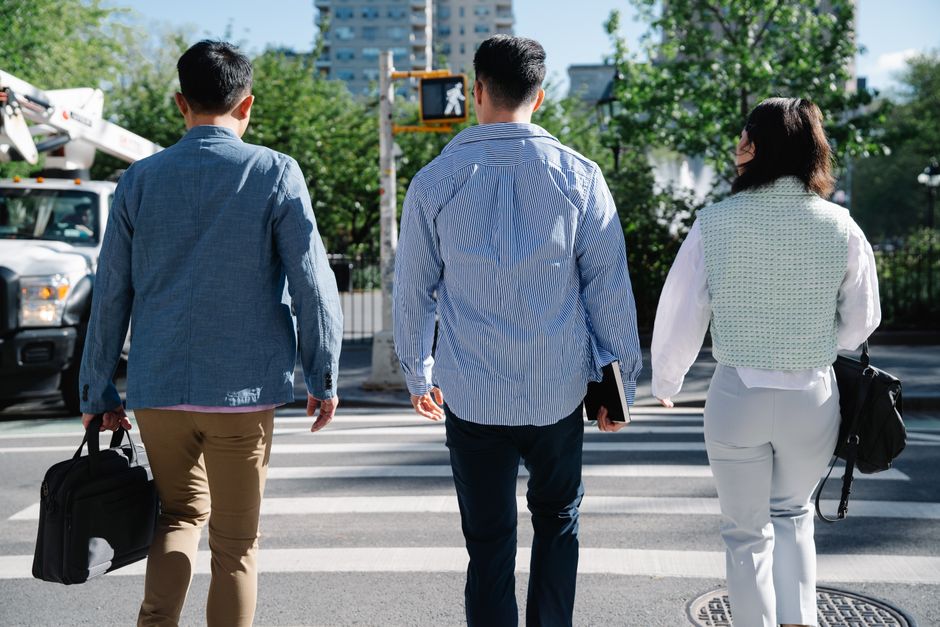 Three adults crossing a city road at a pedestrian crossing, carrying handbags under a clear sky.