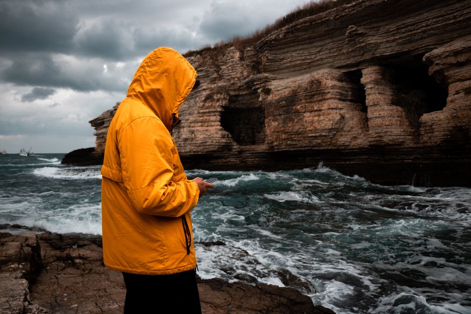 Person in a yellow jacket standing by rocky seashore texting during an overcast day.