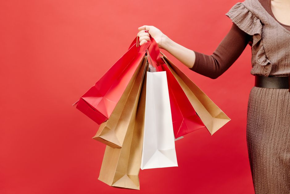 A woman in a brown dress holding multiple shopping bags against a vivid red background.