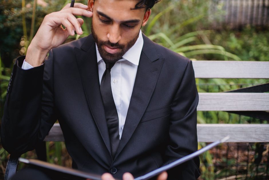 A well-dressed man in a black suit reads intently on an outdoor bench, exuding professionalism.