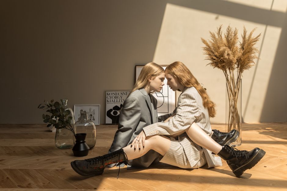 Two women in coats sit intimately on a wooden floor in a studio setting, bathed in natural light and shadows.