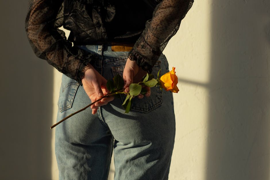 A person wearing jeans holds an orange rose behind their back with soft lighting.