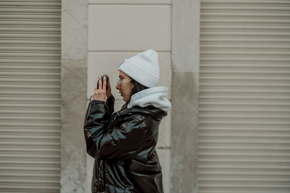 Side view of a woman wearing a beanie and black jacket, holding a camera in an urban envir