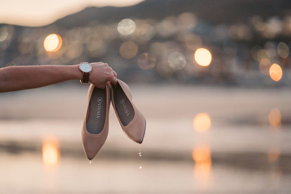 Stylish brown slip-on shoes held by hand at sunset with seaside bokeh background.
