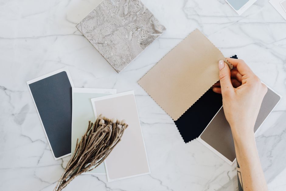 Hands holding fabric swatches with color samples on a marble surface for interior design inspiration.