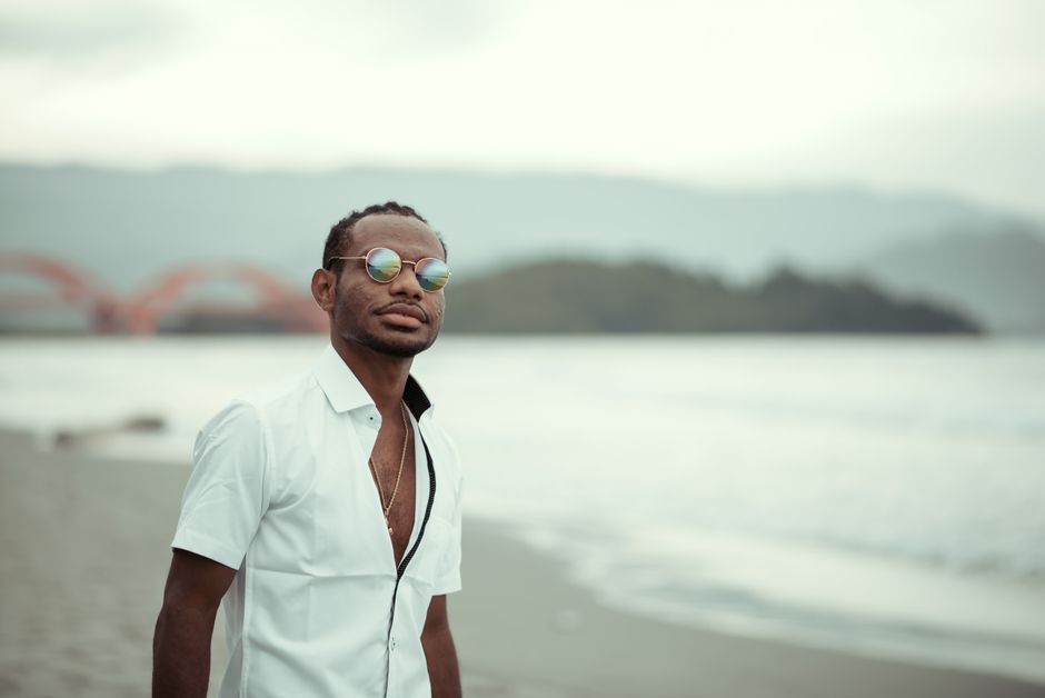 Stylish young man wearing sunglasses, standing on a peaceful beach with distant mountains.