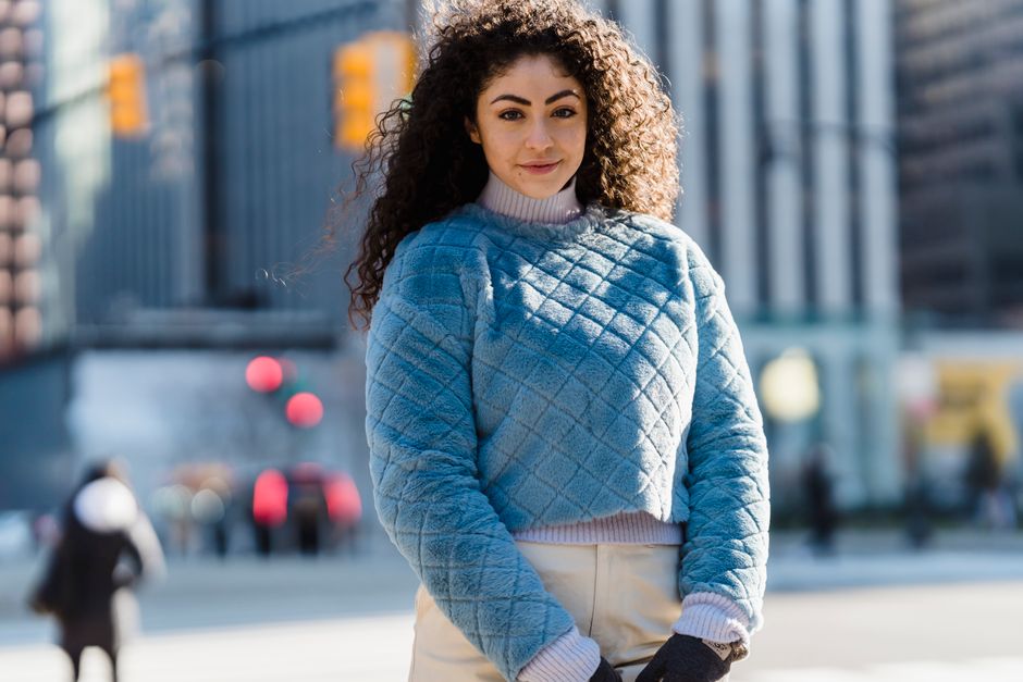 Crop cheerful female with dark curly hair in stylish clothes smiling and looking at camera on blurred background of street