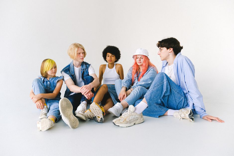 Full body group of thoughtful multiethnic teenager friends in jeans clothes sitting on floor and speaking with each other against white background