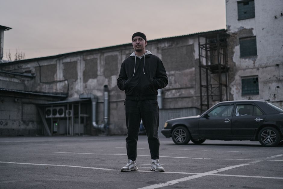 A young man in casual clothing stands in an urban parking lot with a car and industrial backdrop.