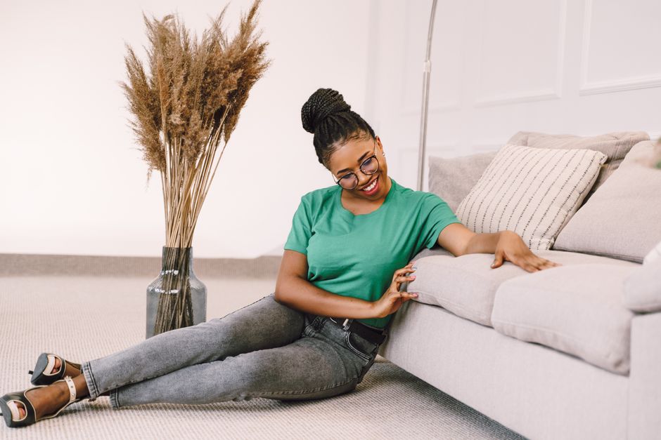 African American woman in casual setting, smiling on the living room floor beside a beige sofa.