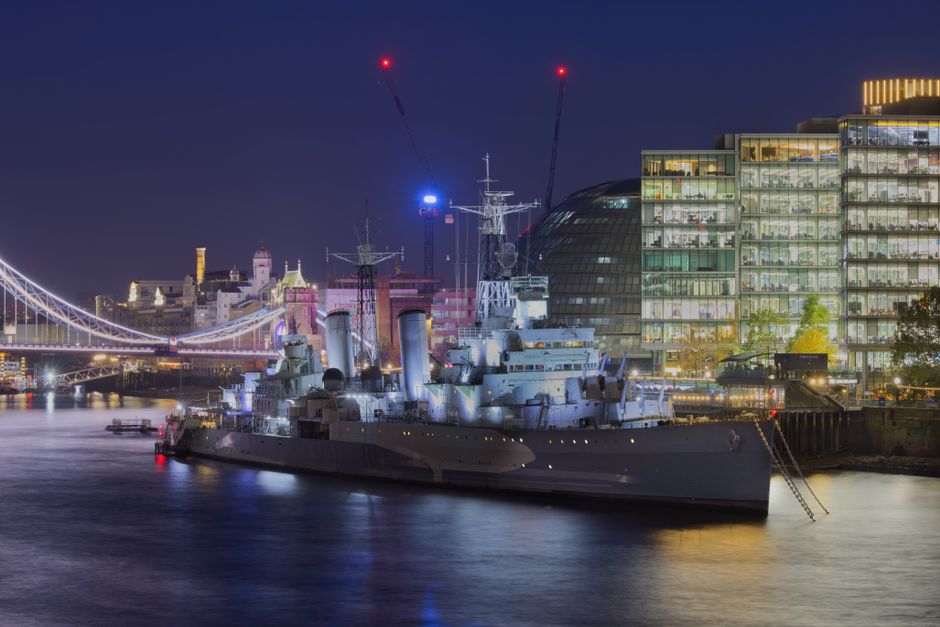 Illuminated HMS Belfast with Tower Bridge in the backdrop at night in London.